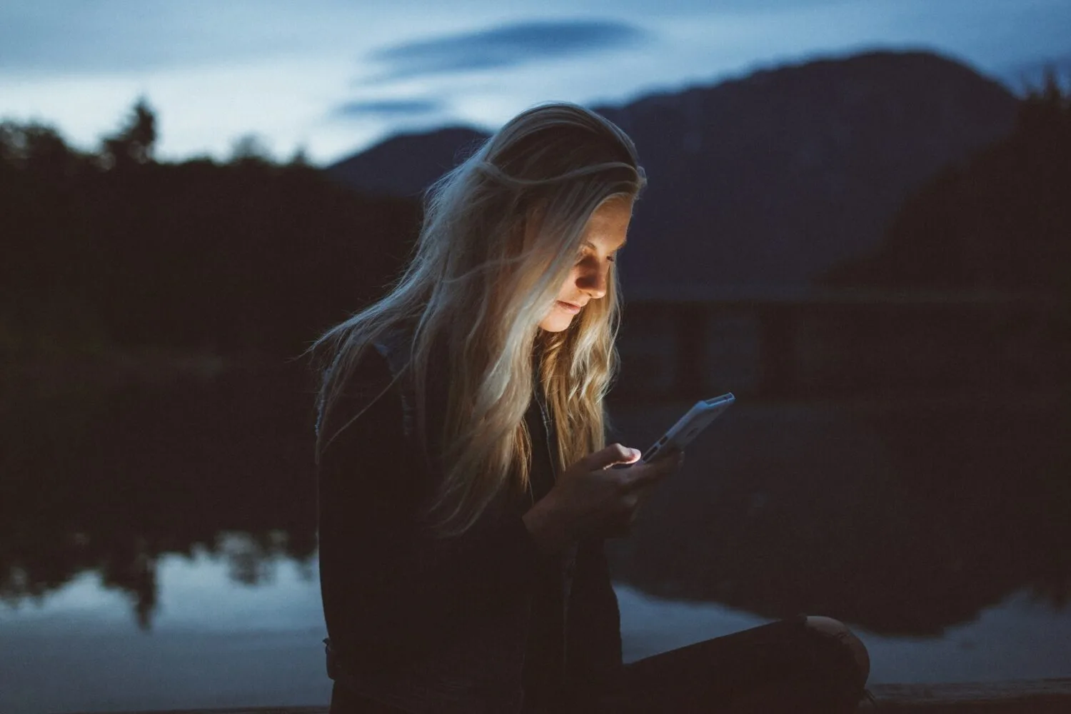 A woman sitting in the dark by a lake looking at her phone