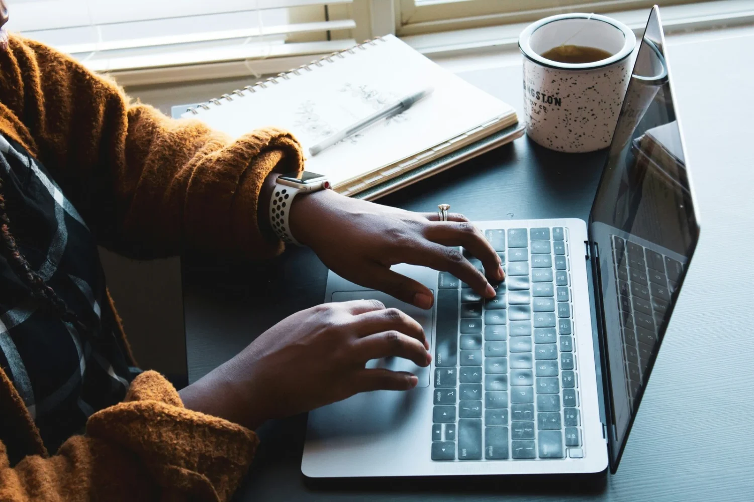 A person typing on a laptop, with a notebook and coffee mug on the desk beside them.