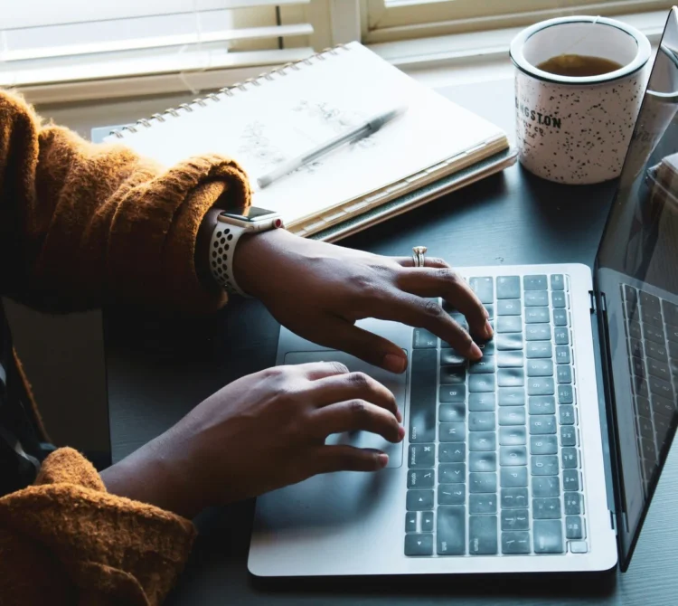 A person typing on a laptop, with a notebook and coffee mug on the desk beside them.