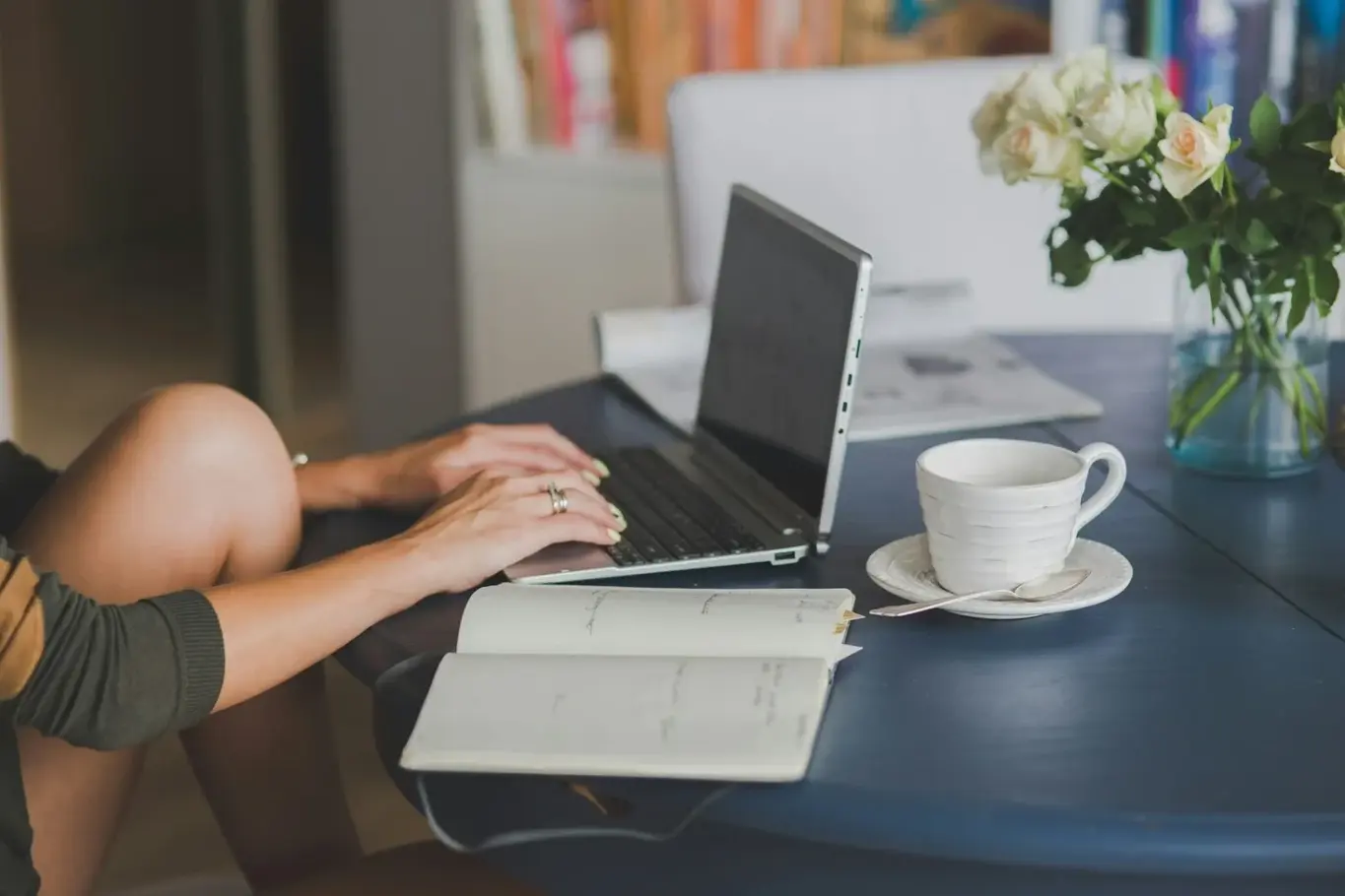 Person using a laptop with an open notebook and a cup of coffee on a table.