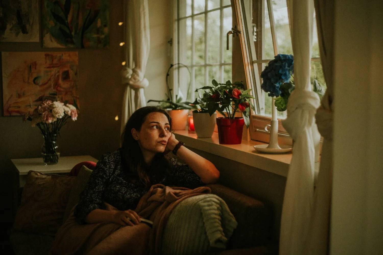 Woman sitting on a couch beside a window in a softly lit room, looking outside.