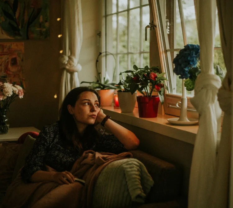 Woman sitting on a couch beside a window in a softly lit room, looking outside.
