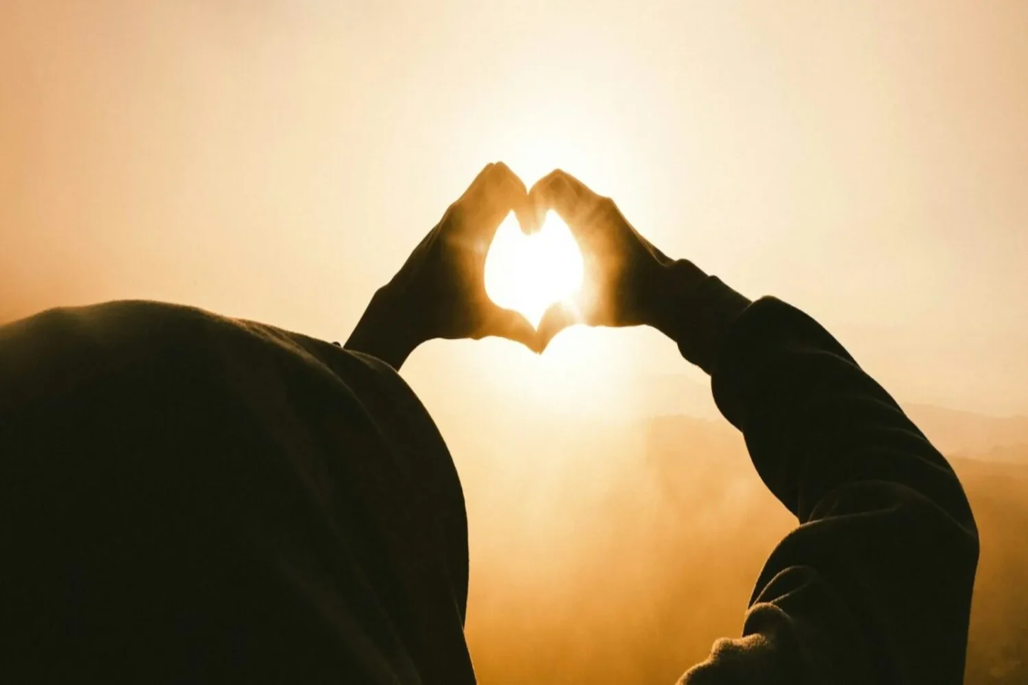 A person making a heart shape with their hands in front of the sun.