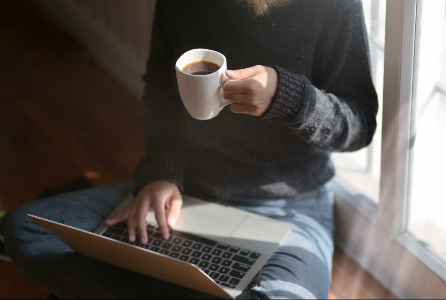 A person holding a cup of tea while sitting crosslegged using a laptop
