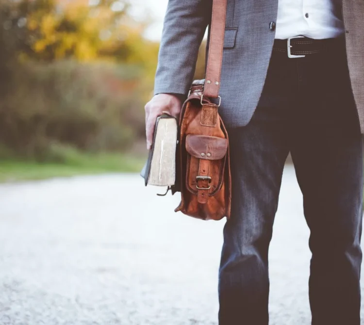 A man wearing a suit holding a book and wearing a brown leather shoulder bag.