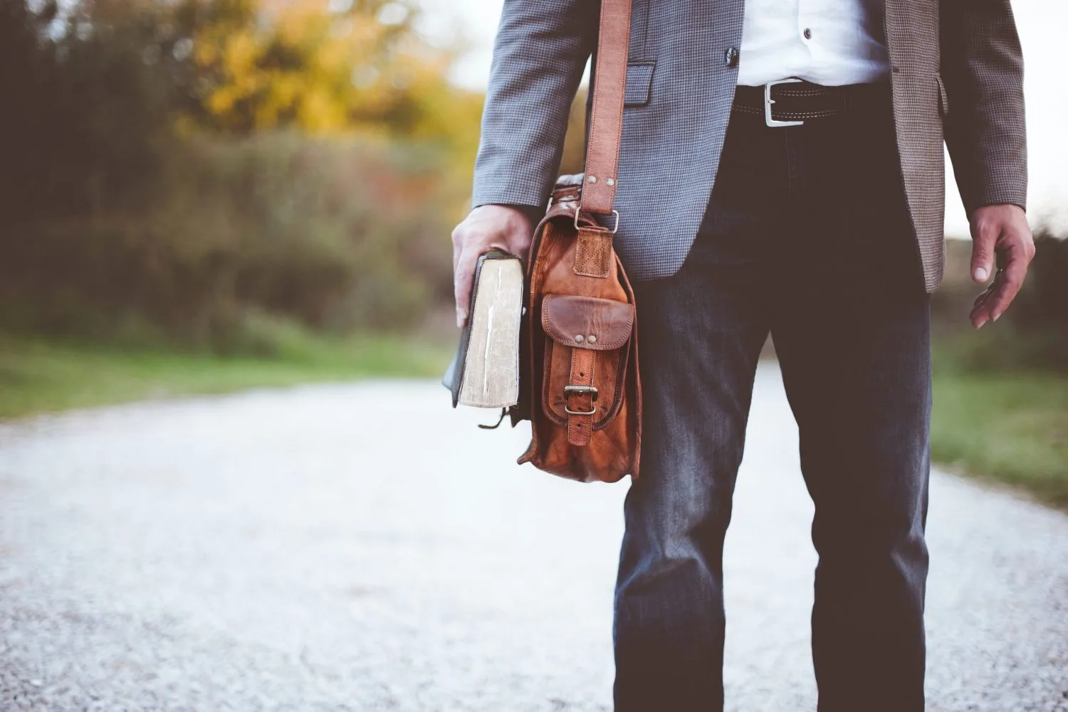 A man wearing a suit holding a book and wearing a brown leather shoulder bag.