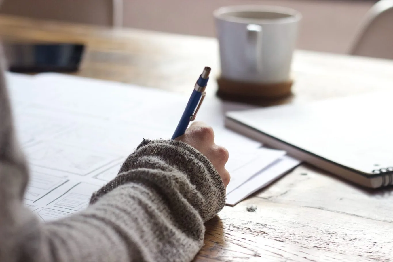 A person wearing a knitted sweater writing on a sheet of paper with a blue pen at a wooden table.