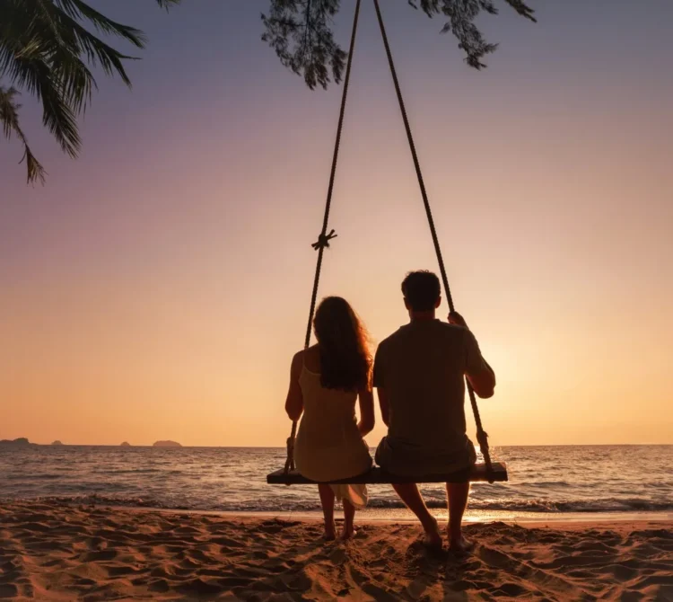 Two people sitting on a swing on a beach in front of the sunset