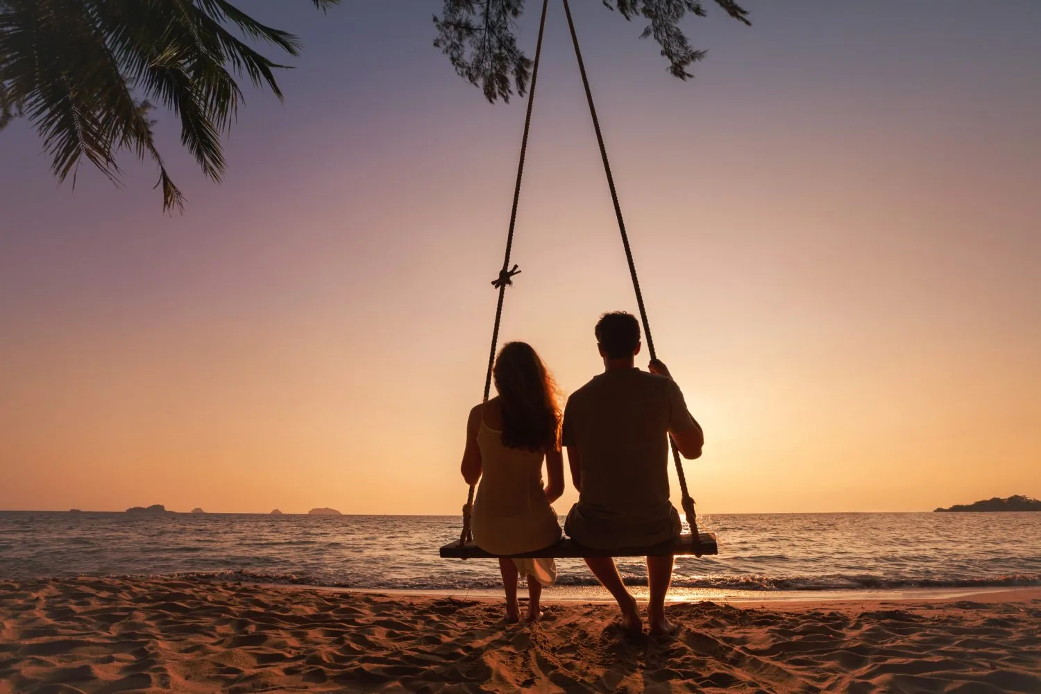 Two people sitting on a swing on a beach in front of the sunset