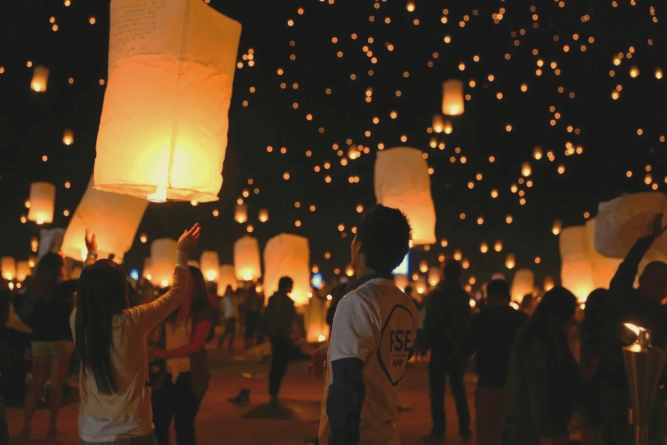 People releasing illuminated lanterns into the night sky during an outdoor gathering.