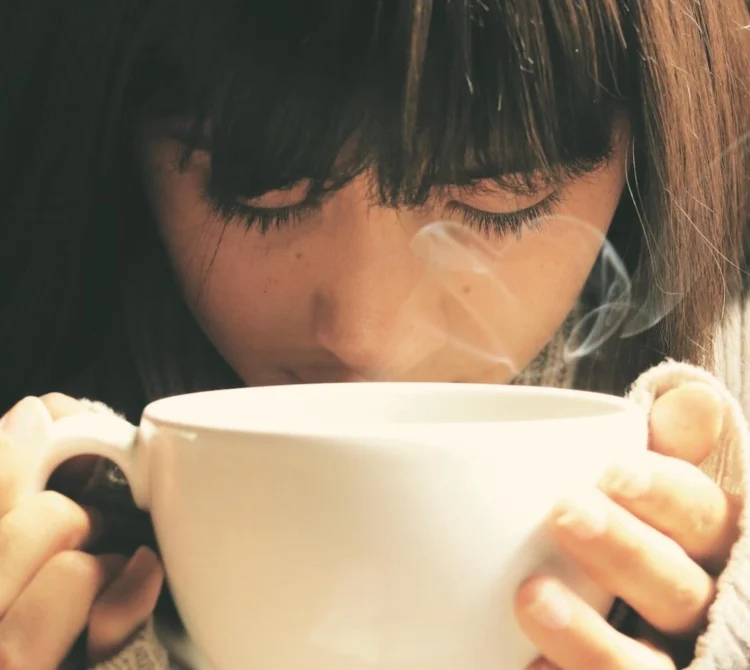 A woman with her eyes closed holding a warm drink.