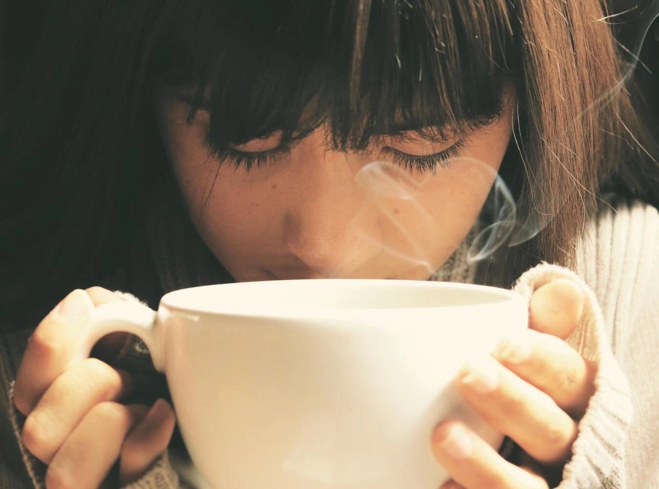 A woman with her eyes closed holding a warm drink.