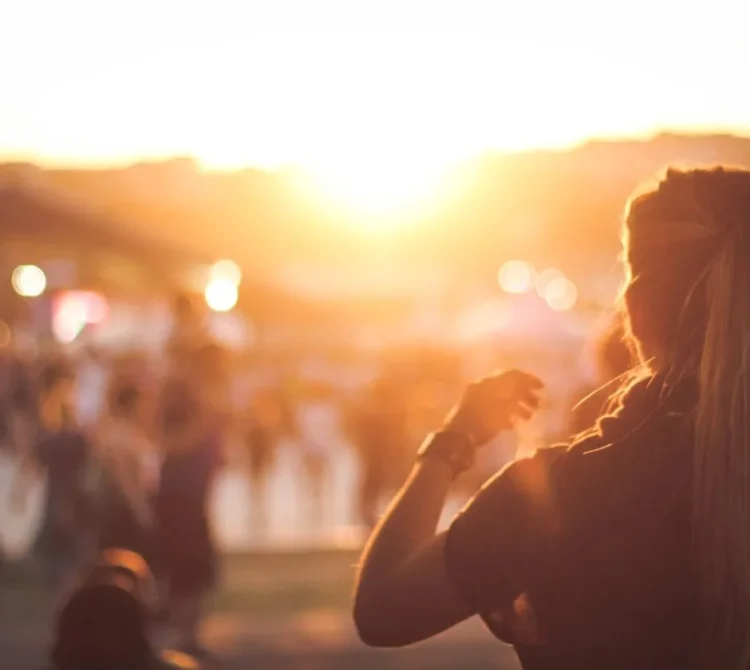 A woman standing outdoors facing the sunset with people in the background.