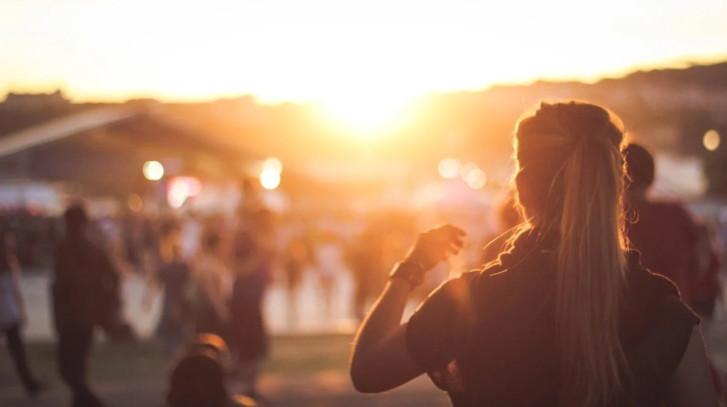 A woman standing outdoors facing the sunset with people in the background.