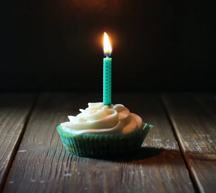 A cupcake with white frosting and a single lit birthday candle on a wooden table.