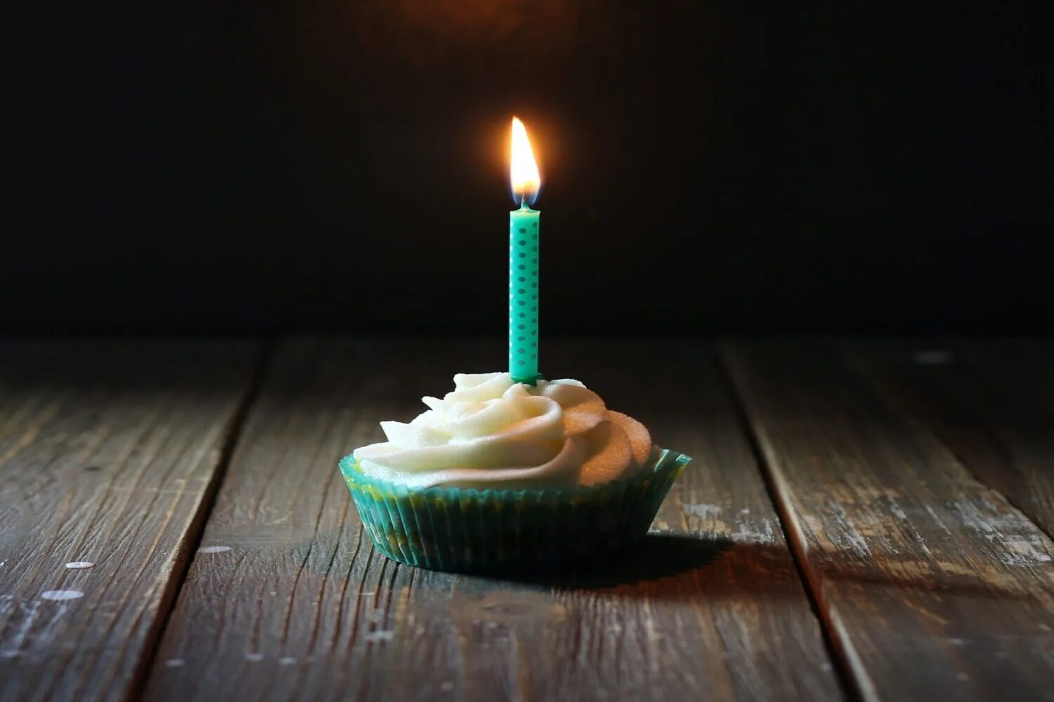 A cupcake with white frosting and a single lit birthday candle on a wooden table.
