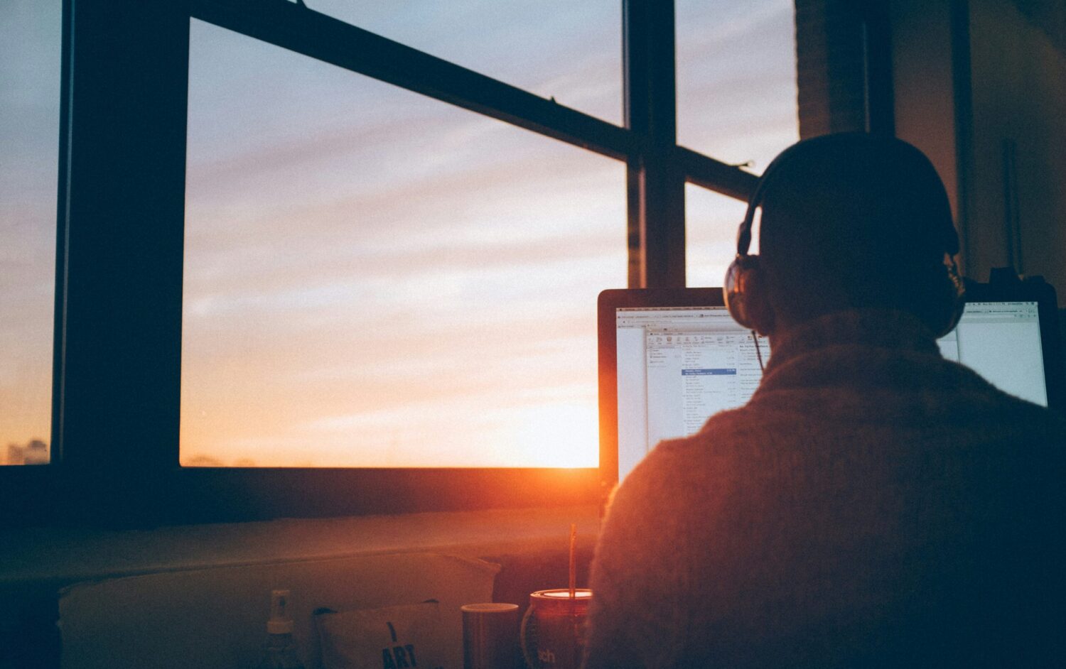 Person wearing headphones facing a computer monitor in front of a window showing setting sun