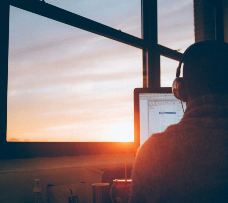 Person wearing headphones facing a computer monitor in front of a window showing setting sun