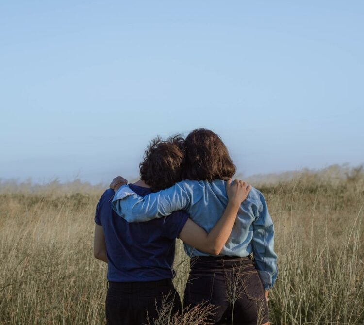 Two people stand in a field facing away from the camera with their arms around each others' shoulders