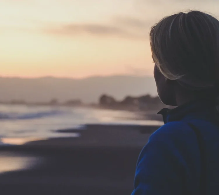 A woman looking toward the beach and ocean at sunset with mountains in the distance.