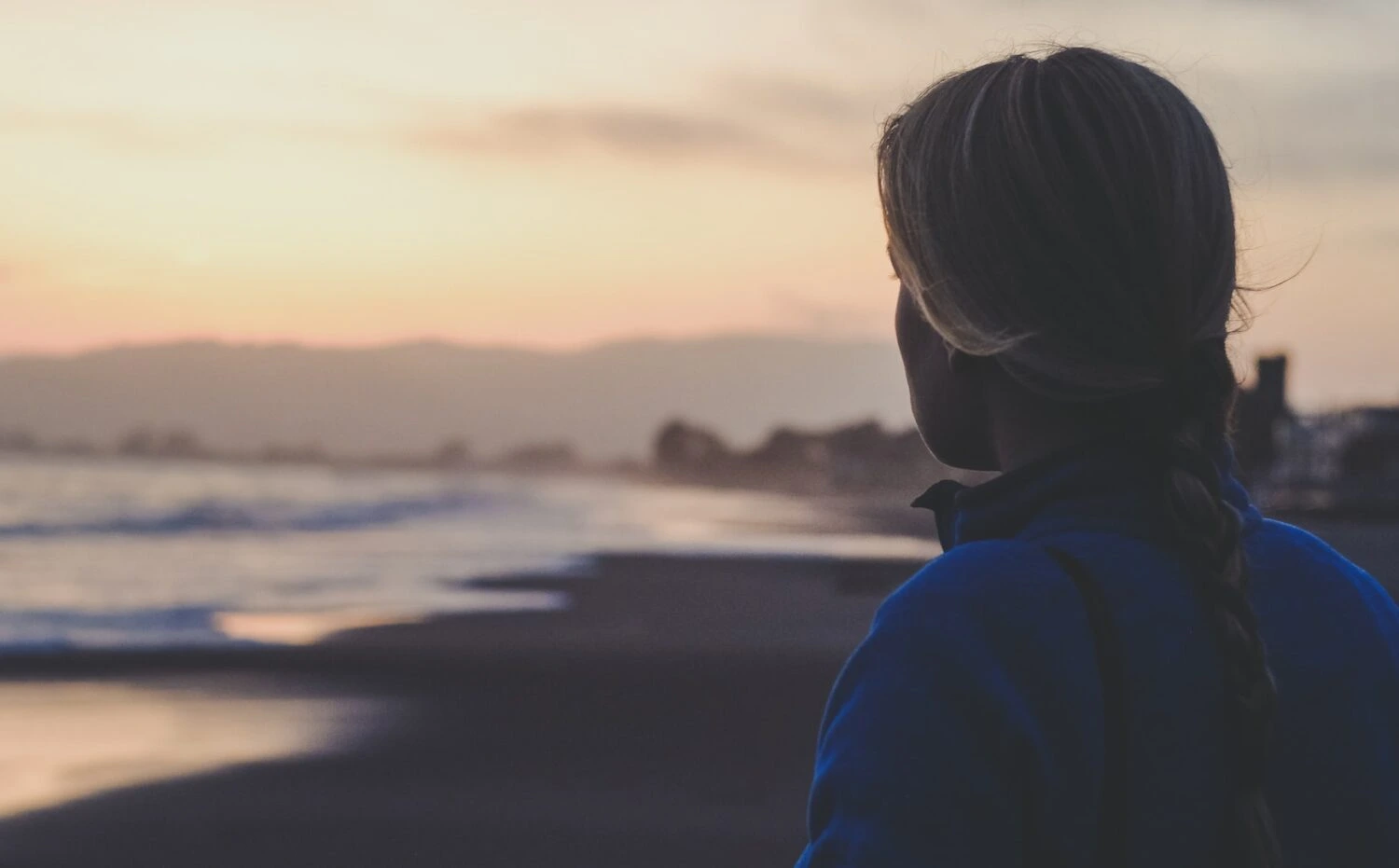 A woman looking toward the beach and ocean at sunset with mountains in the distance.
