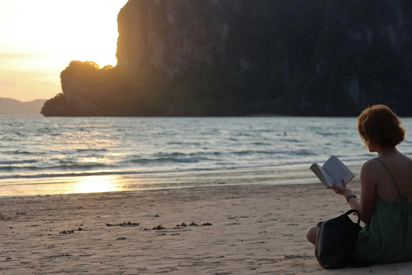 A woman sitting on the beach reading a book while looking out over the water at sunset.