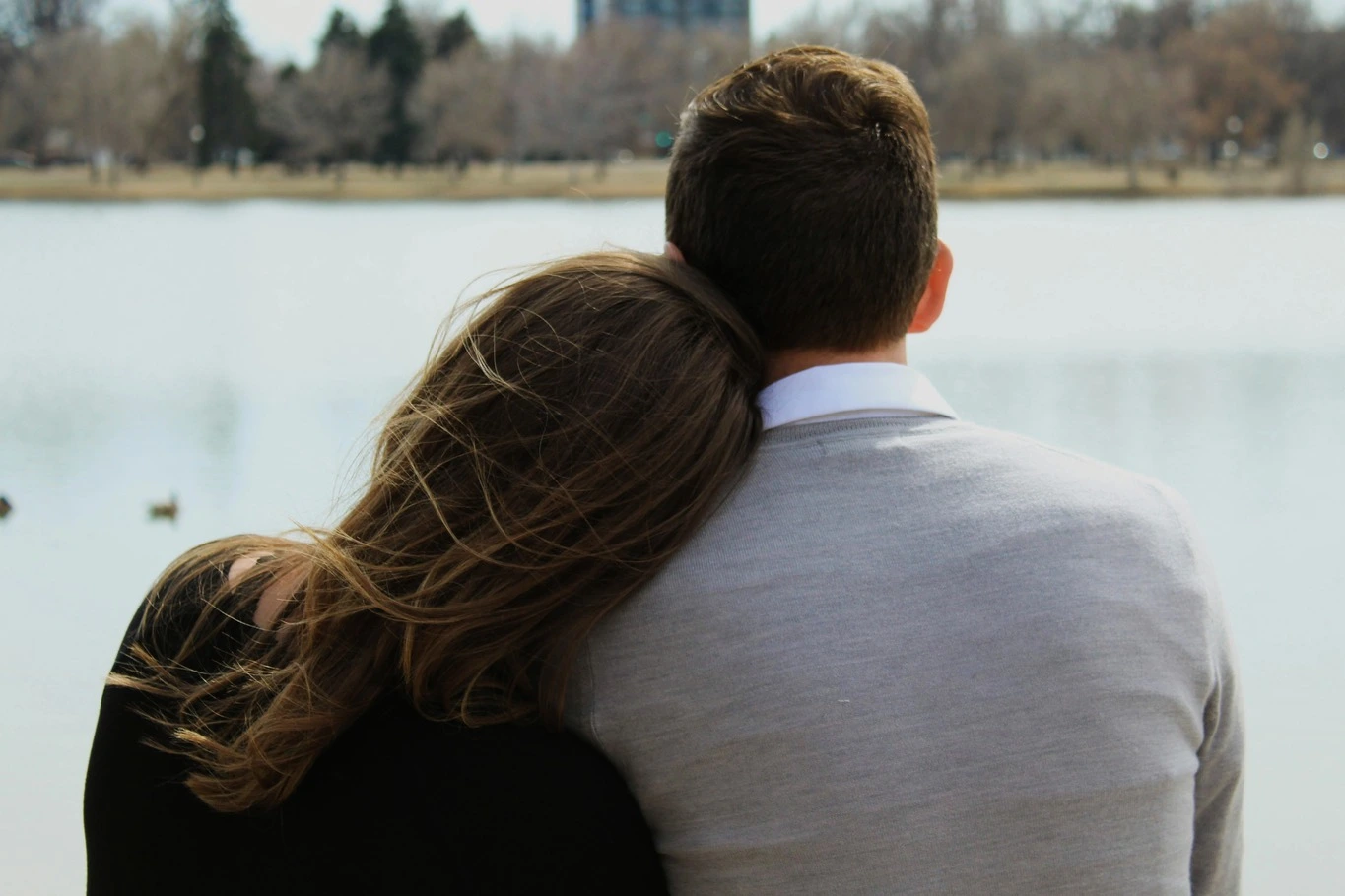 Couple sitting beside a lake with one person resting their head on the other's shoulder.