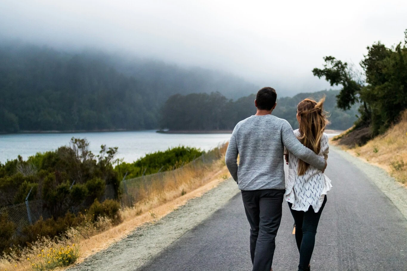 A couple walking side by side on a lakeside road with one person wrapping an arm around the other's shoulders.