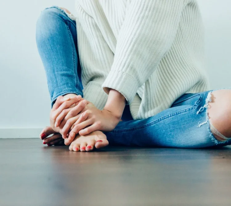A person sitting cross-legged on the floor wearing a sweater and blue jeans.