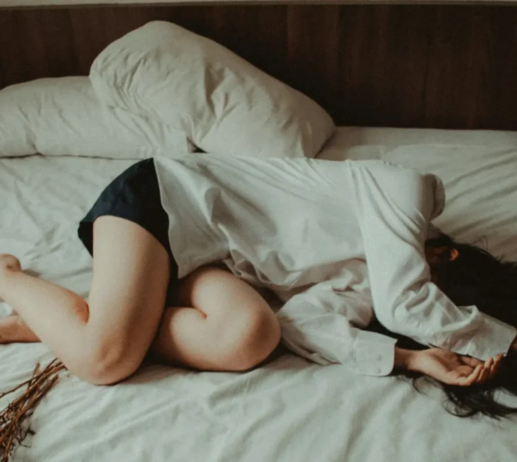 A woman lying on a bed in a fetal position with her hands covering her face.