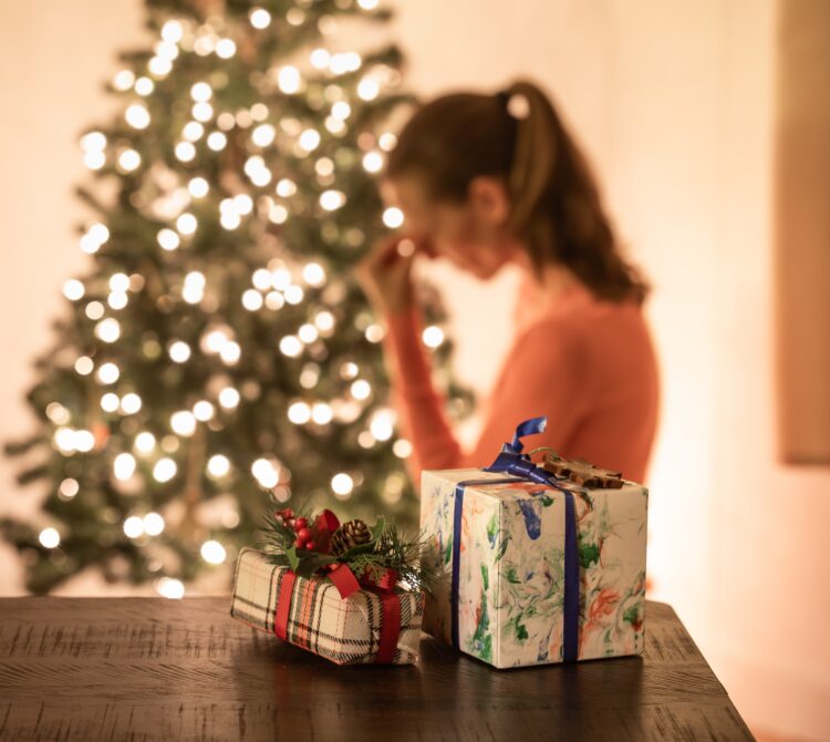 A woman with her head in her hands, standing in front of a Christmas tree with presents