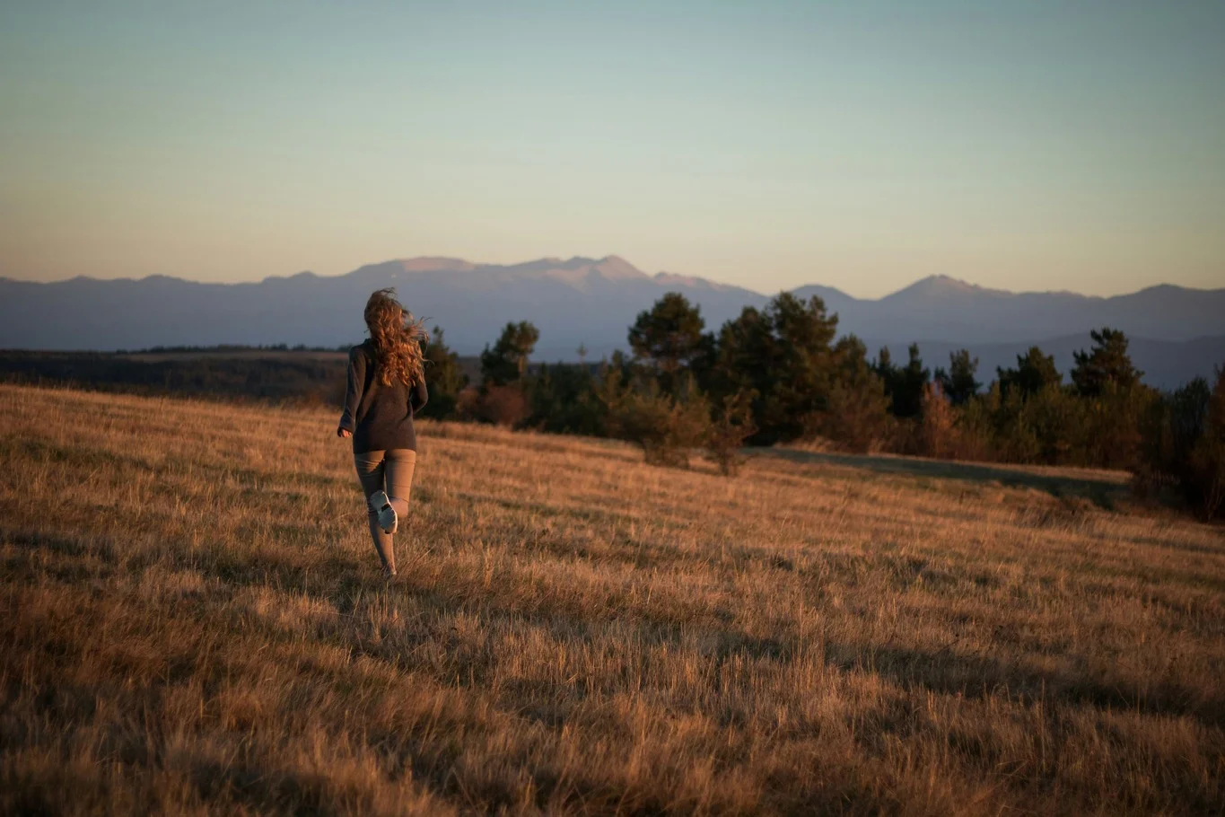 A woman running across an open field toward distant mountains at sunset.