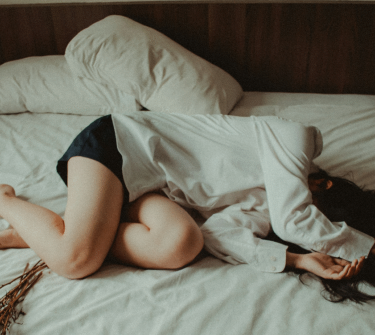Women lying on bed white sheets in fetal position hands covering face