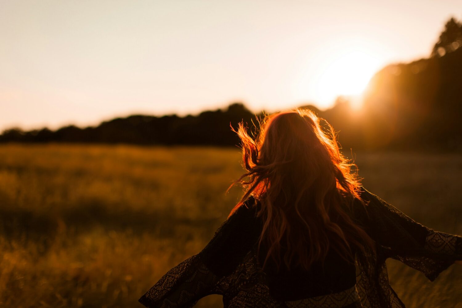A woman in a paddock facing away from the camera, towards a sunset, arms open wide