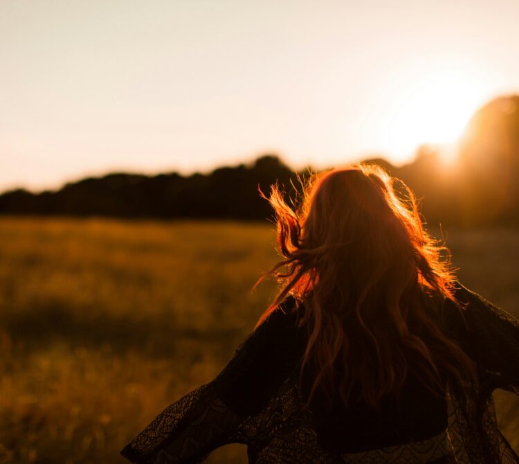 A woman in a paddock facing away from the camera, towards a sunset, arms open wide