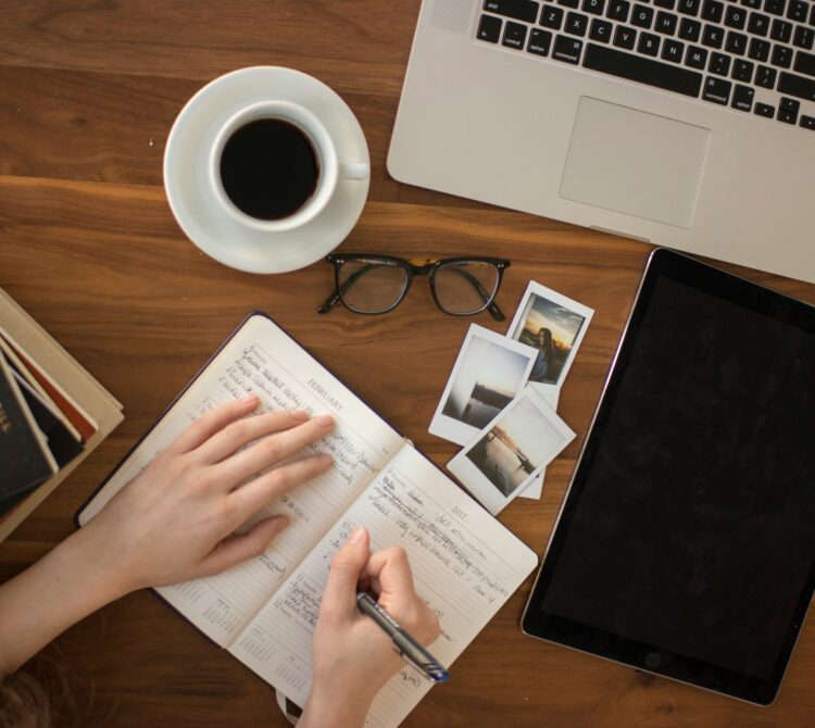 Work desk with laptop, cup of coffee, glasses, photos and hands writing in a diary/planner