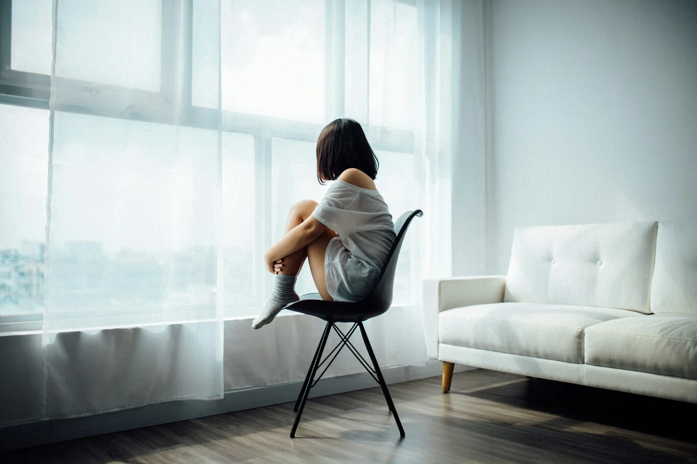 A person sitting on a chair by a window with their knees pulled up.
