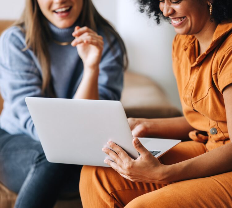 two women sitting on a couch looking at a computer smiling