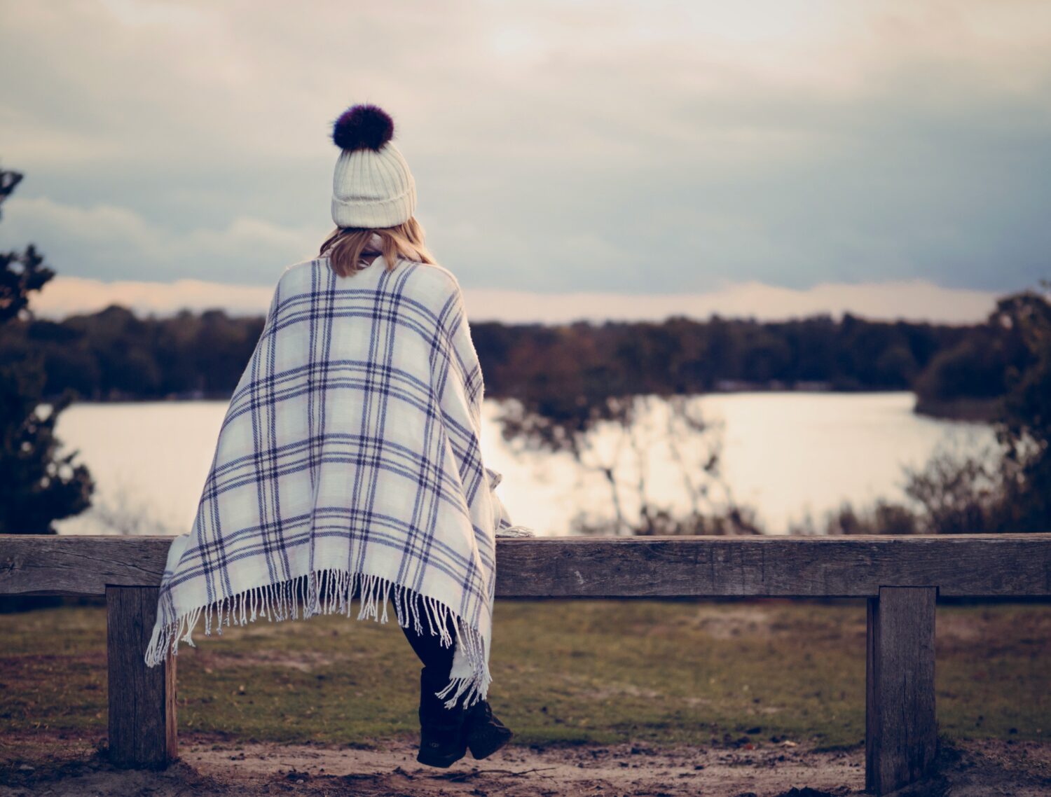 Person in beanie sitting on bench looking out at lake of water