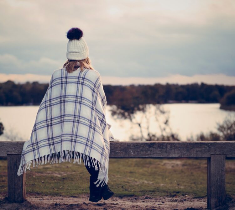 Person in beanie sitting on bench looking out at lake of water