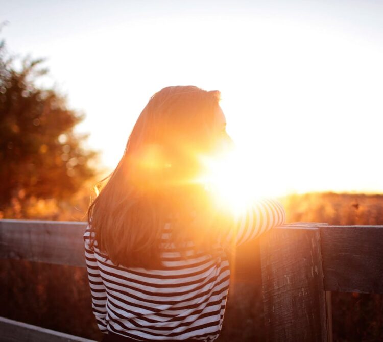 A woman standing outside, leaning on a fence, looking at the sunset