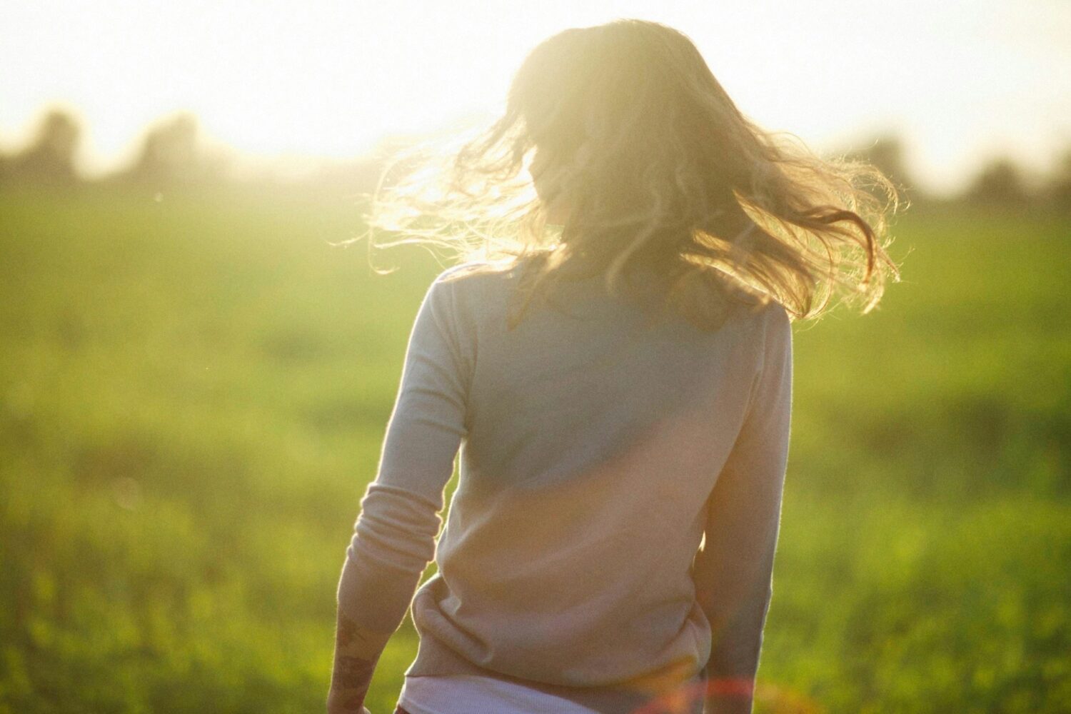 A woman with hair let out, standing facing away from the camera in a green paddock
