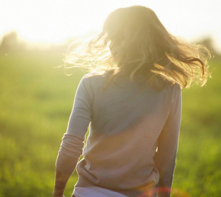 A woman with hair let out, standing facing away from the camera in a green paddock
