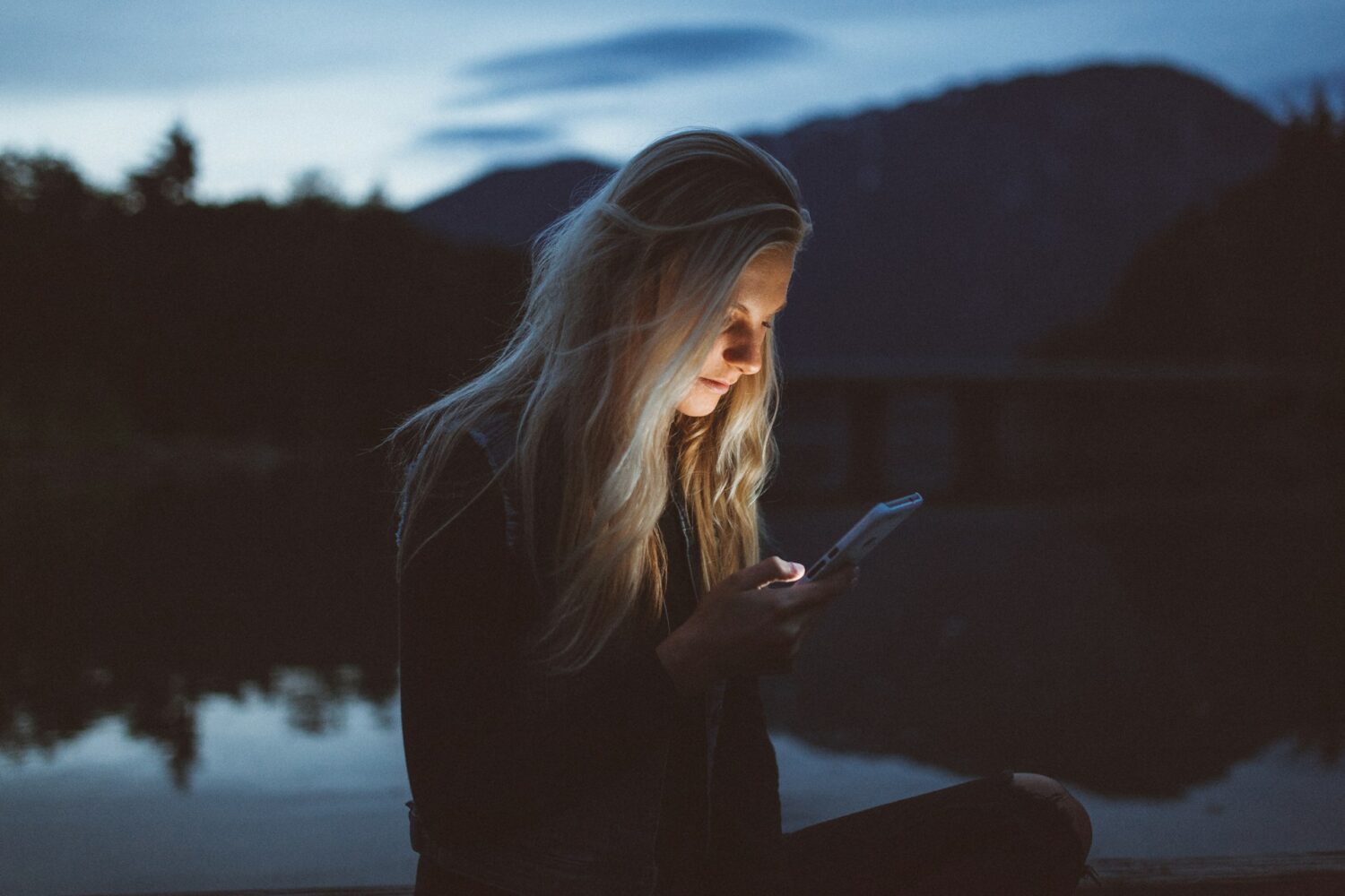 A woman sitting in the dark, staring at her phone, her face lit up by the phone's light