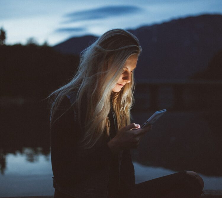 A woman sitting in the dark, staring at her phone, her face lit up by the phone's light