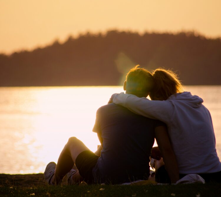 Two people sitting by lake durning sunset with arms wrapped around one another