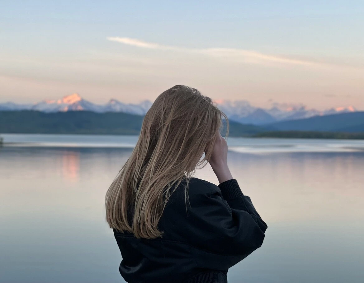 A woman standing by a calm lake looking toward distant mountains.