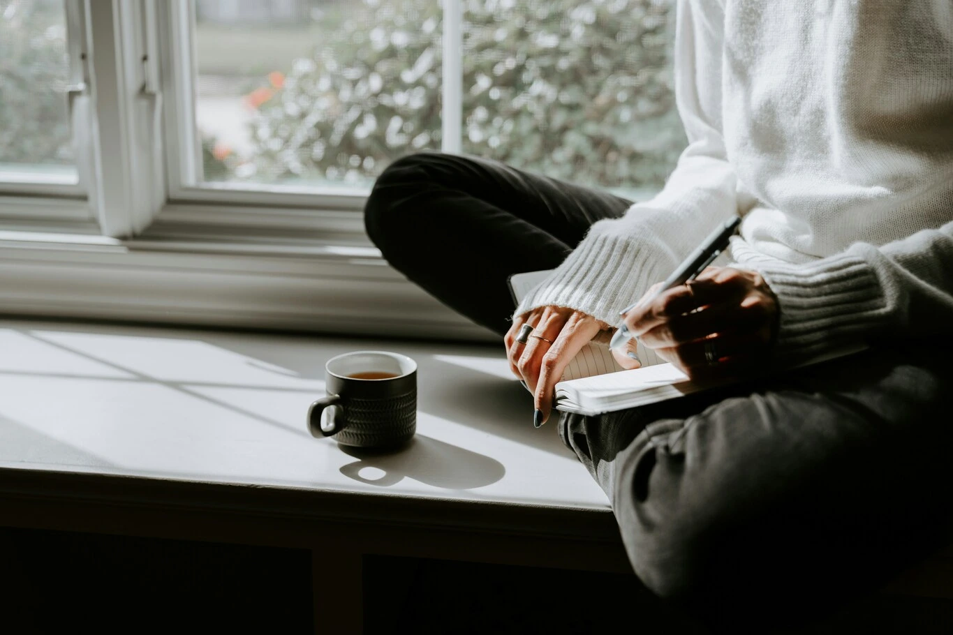 A person writing in a journal while sitting beside a window with a mug nearby.