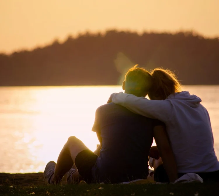 Two people sitting by lake durning sunset with arms wrapped around one another