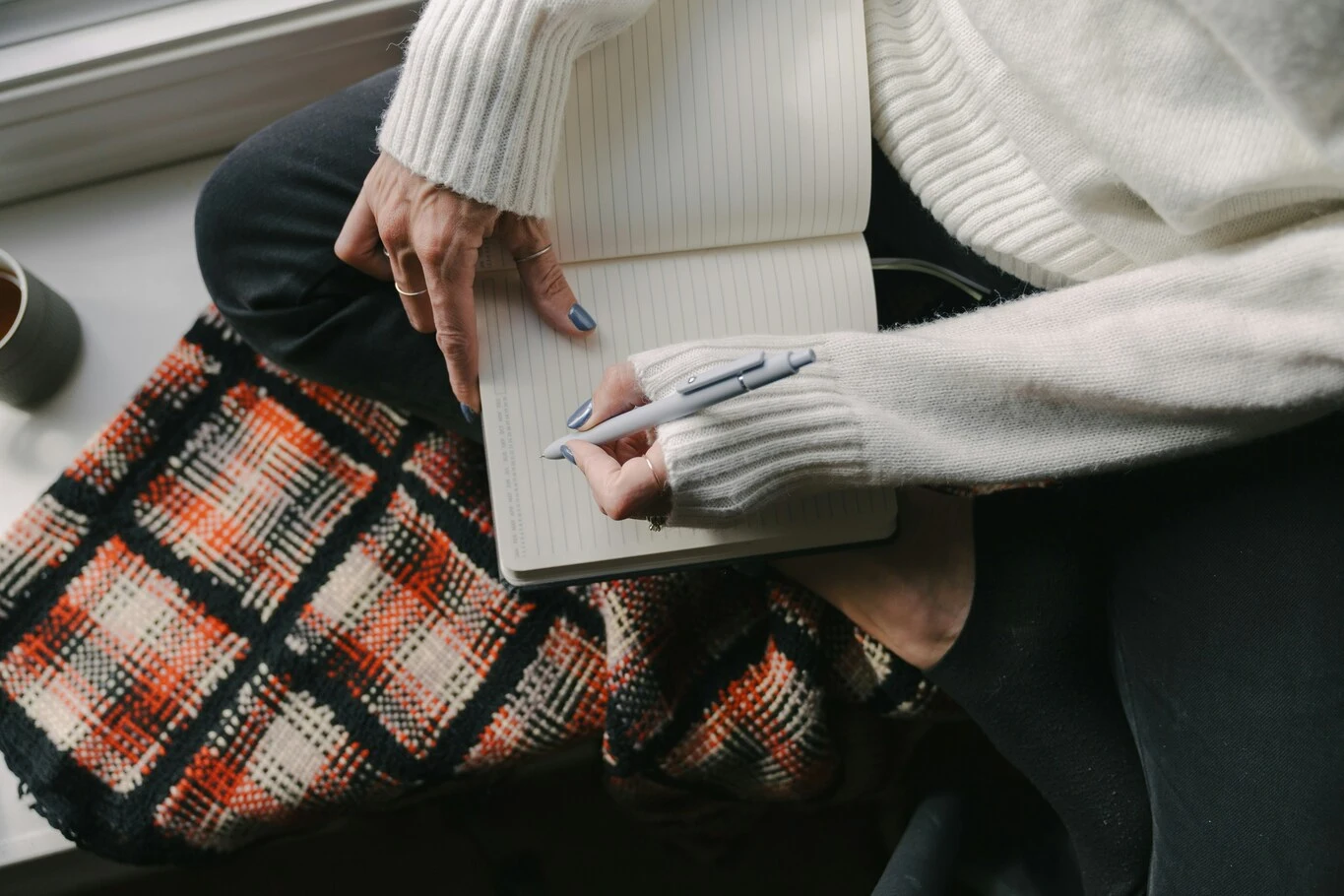 person writing in an open notebook while sitting beside a window